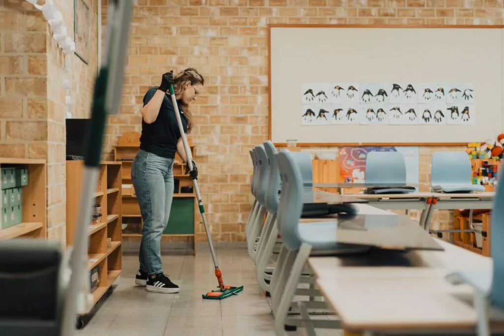 Three professional cleaners in green and red overalls working in a modern kitchen, mopping the floor, and cleaning high walls and windows.