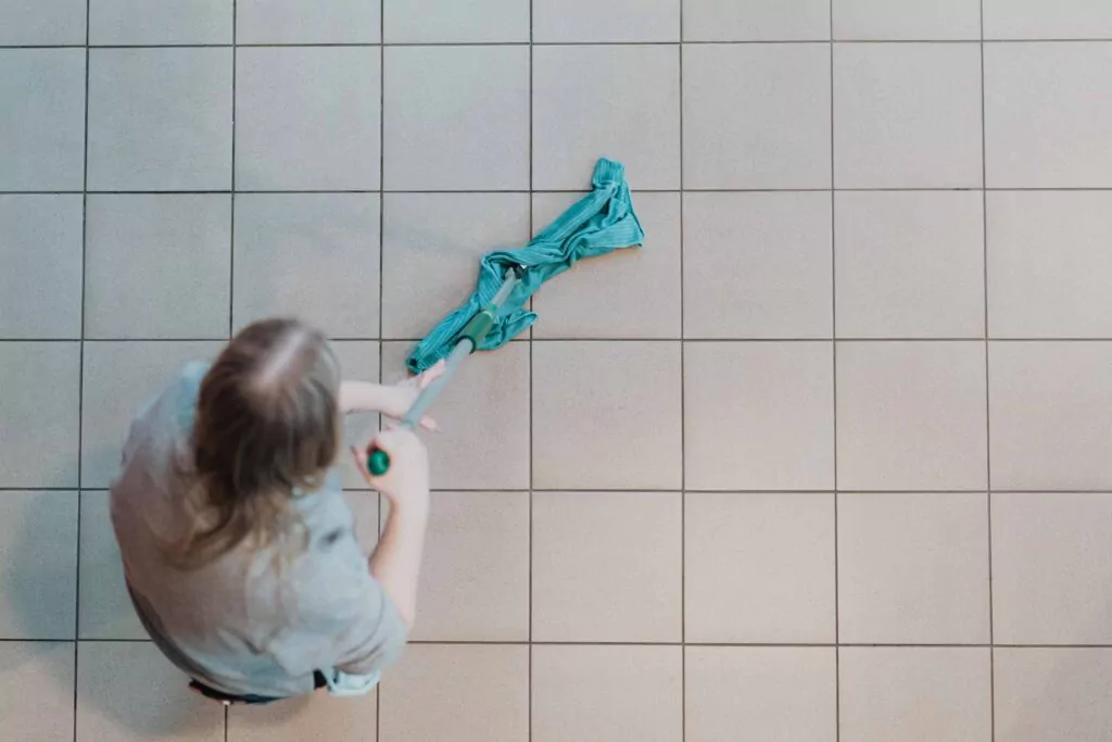 Three professional cleaners in green and red overalls working in a modern kitchen, mopping the floor, and cleaning high walls and windows.