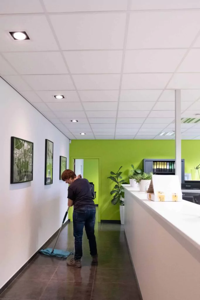 Three professional cleaners in green and red overalls working in a modern kitchen, mopping the floor, and cleaning high walls and windows.